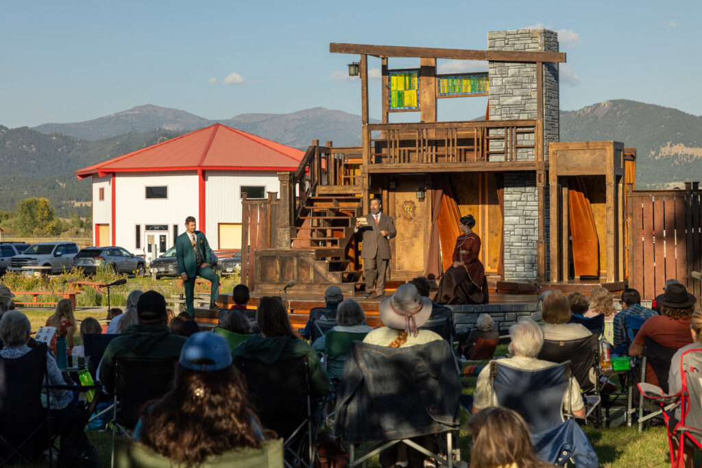 Live performance of Montana Shakespeare in the Park in Boulder, Montana. Crowd of people stands in front of a stage watching the actors in a free, open air showing of Shakespeare.