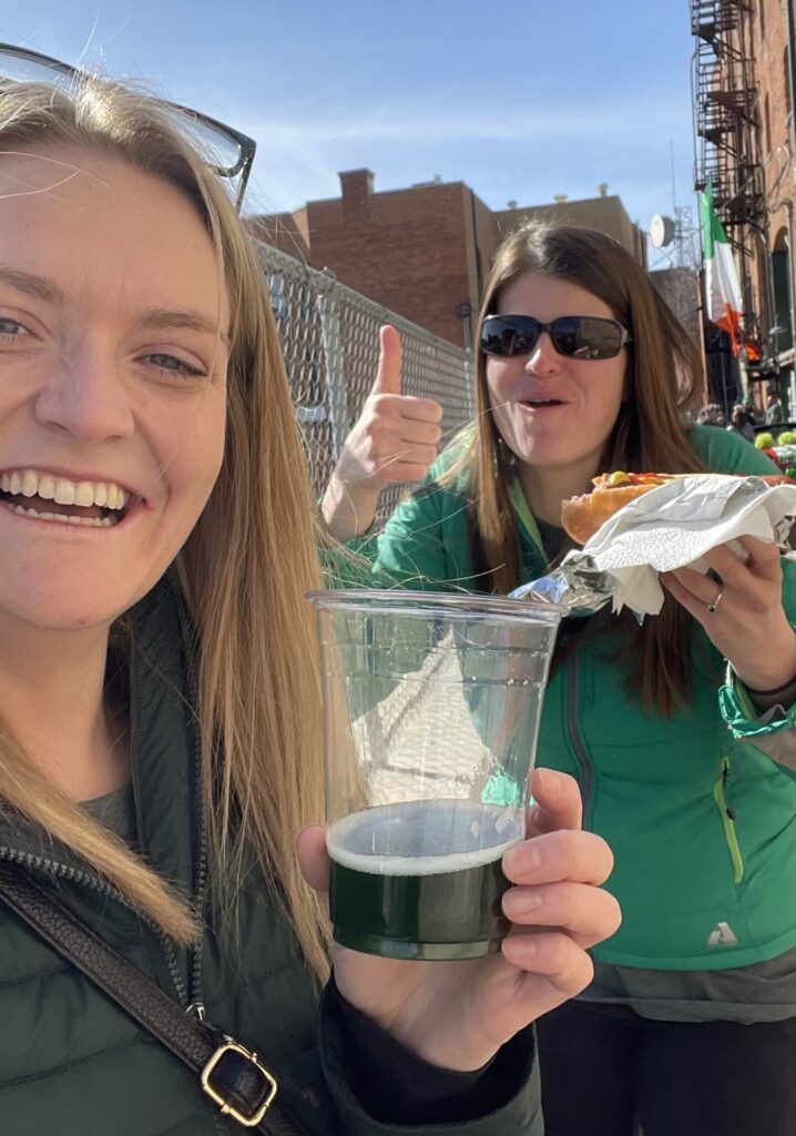 Two women laugh and give a thumbs up while celebrating St. Patrick's Day in Uptown Butte, Montana, holding a green beer from Quarry Brewing and a hot dog from Terminal Meat Market, with an Irish flag flying in the background along the historic Uptown Butte streetscape. Photo by Tempest Technologies