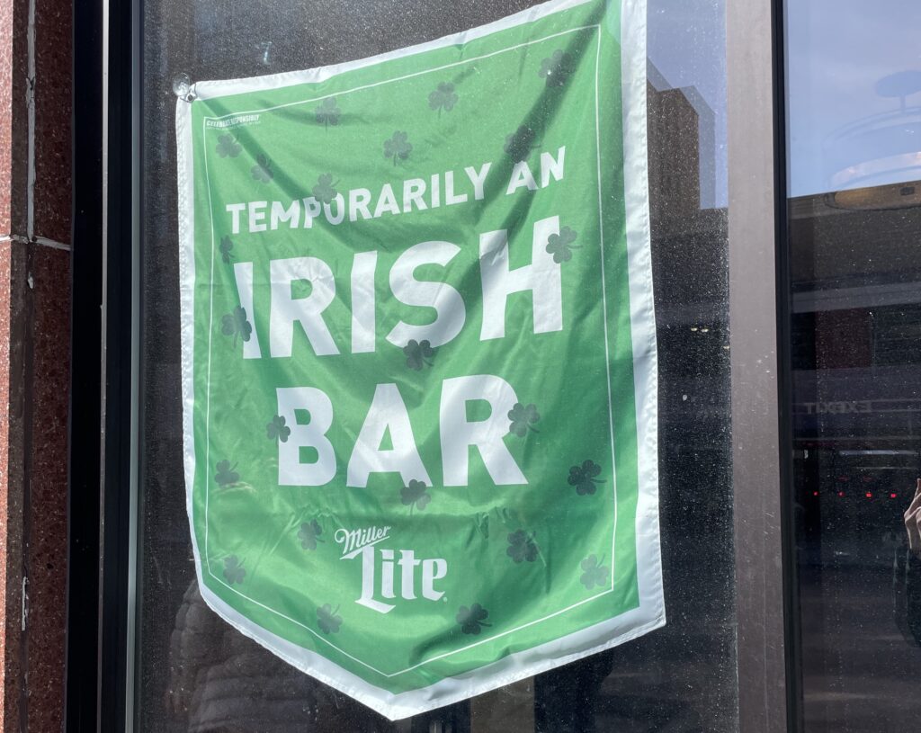 A green shamrock-covered Miller Lite flag reading "Temporarily an Irish Bar" hangs in the window of a bar along the St. Patrick's Day parade route in Uptown Butte, Montana — a nod to the city's Irish heritage and one of the most celebrated St. Patrick's Day destinations in the United States. Photo by Tempest Technologies.