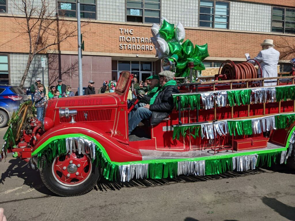 A vintage red fire truck decorated with green and silver fringe and shamrock balloons rolls through Uptown Butte during the annual St. Patrick's Day Parade in Butte, Montana, with spectators lining the street in front of The Montana Standard building. Photo by Tempest Technologies