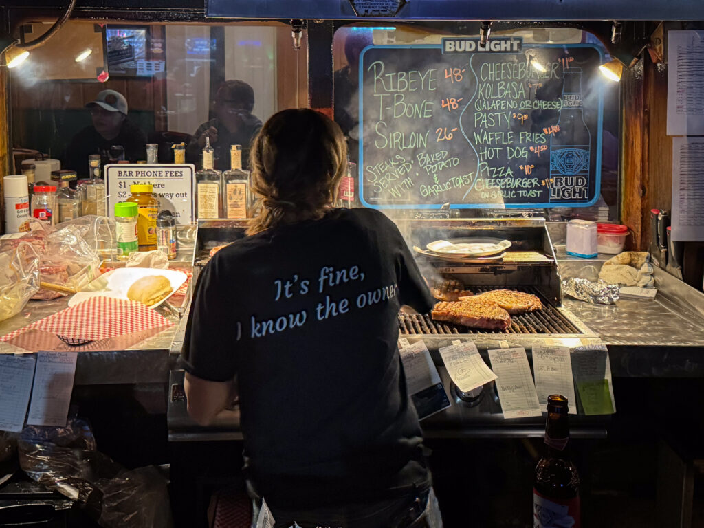 Someone is searing hand-cut steaks on a small grill behind the bar at the Windsor Bar in Boulder, Montana. Located on Main Street in Boulder, MT, the Windsor Bar is a local hangout spot serving hand-cut steaks, baked potatoes, wine, cold beer, and cocktails. The Windsor is famous for its small grill, located right behind the bar, where you can watch your steak being cooked right in front of you.