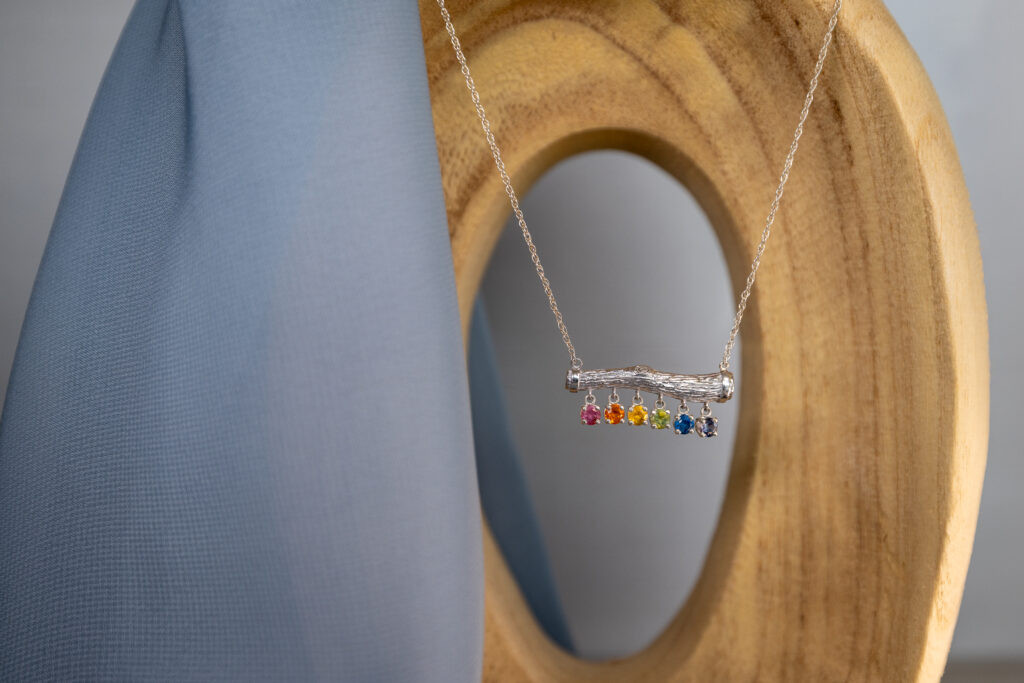 Sterling silver necklace with a twig-style bar pendant and six dangling Montana sapphires in a rainbow of colors (pink, orange, yellow, green, blue, purple), displayed on light wood with a soft blue fabric backdrop. Ethical Montana sapphire jewelry gift.