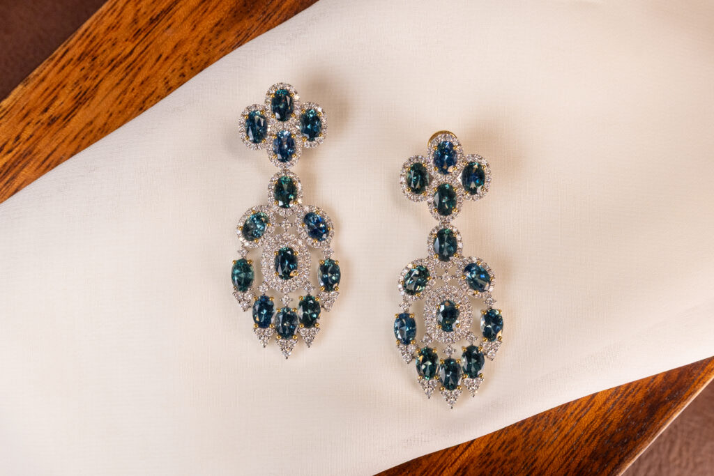 Statement chandelier earrings featuring deep blue Montana sapphires set in oval clusters and framed with sparkling diamonds, displayed on soft ivory fabric and warm wood. Ethical Montana sapphire fine jewelry, elegant luxury gift idea.