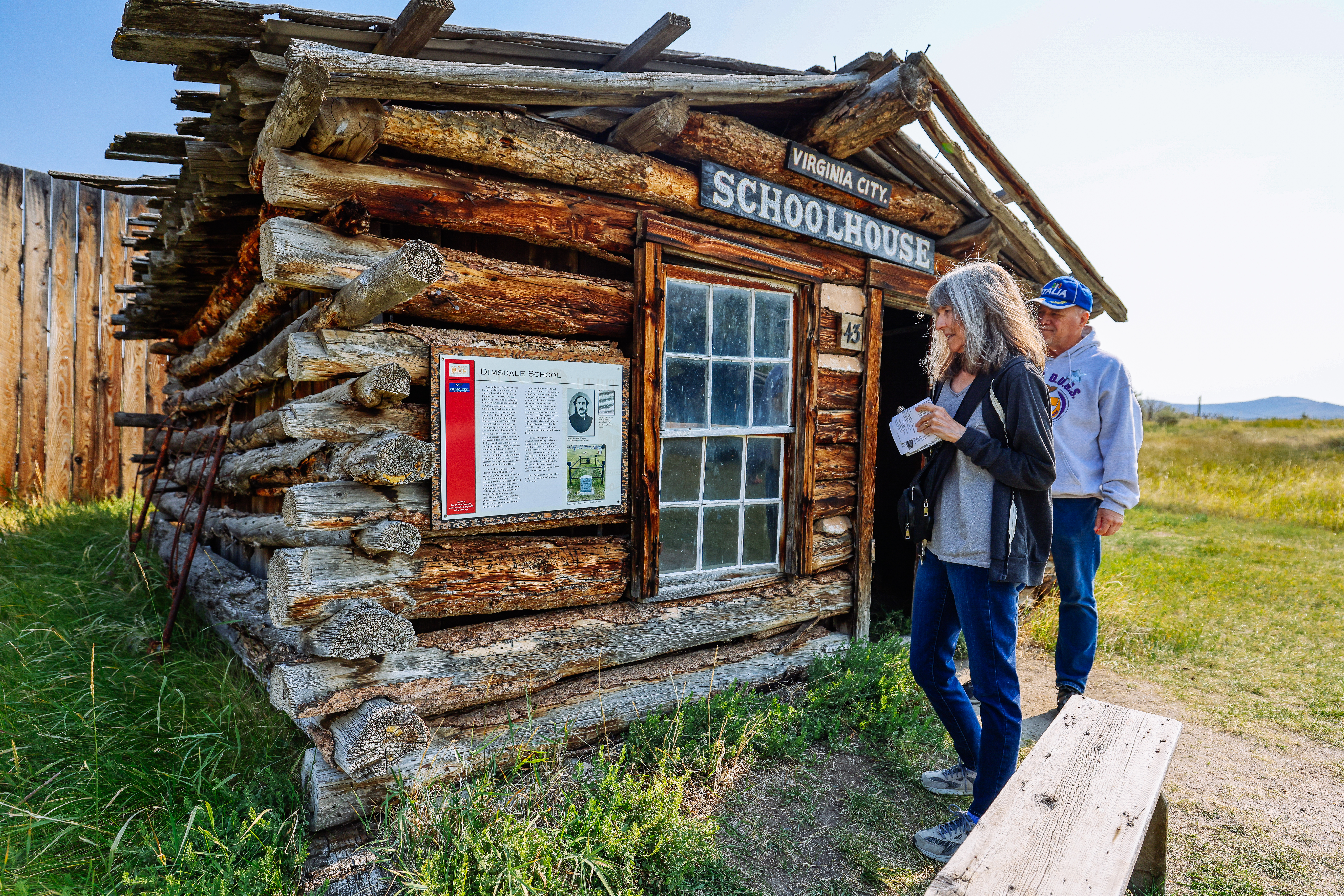 Two people are reading an interpretive sign outside of the original 1860s Virginia City schoolhouse in Virginia City, Montana. An original 1860s gold rush town and the territorial capital of Montana has been preserved and is now a ghost town with museums, living history, train rides, old old-fashioned ice cream store, vaudeville plays, and more.