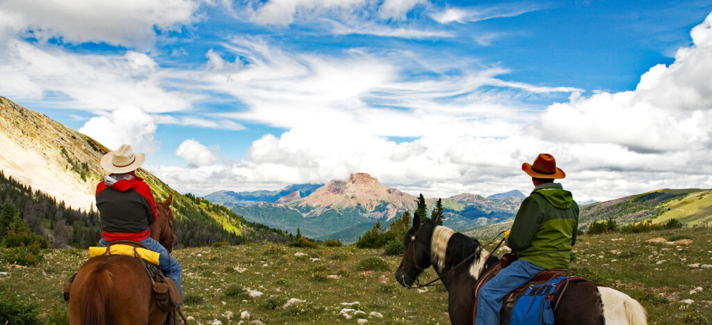Two people on horseback wearing cowboy hats are facing the horizon in front of them, surrounded by the dramatic and gorgeous Madison Mountain Range in Southwest Montana.