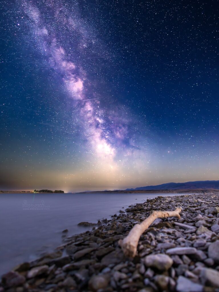A star-filled night sky and bright Milky Way arch over Canyon Ferry Reservoir near Helena, Montana, viewed from a rocky beach with driftwood and still water leading toward distant hills. Photo by Elbie Wood Photography on Instagram.