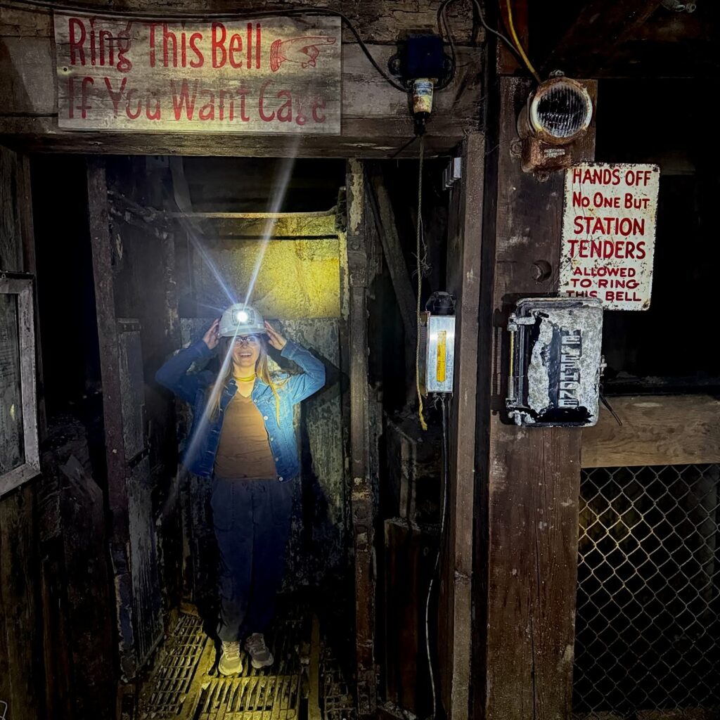 A visitor in a hard hat with a bright headlamp stands inside the wooden, weathered mine elevator at the Orphan Girl Mine on the underground tour at the World Museum of Mining in Butte, Montana, surrounded by vintage signs and mining equipment. Photo submitted by @everywherewitheva on Instagram.