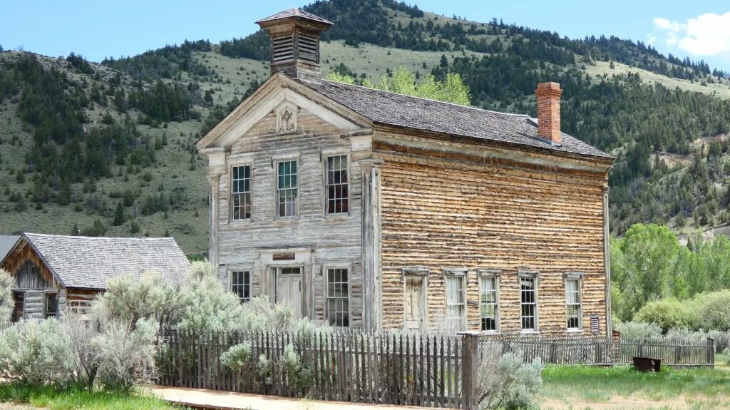The old two-story wooden schoolhouse at Bannack State Park near Dillon, Montana, a preserved ghost town, sits behind a picket fence with sagebrush in front and green hills rising in the background. Photo by @guliestrip on Instagram.