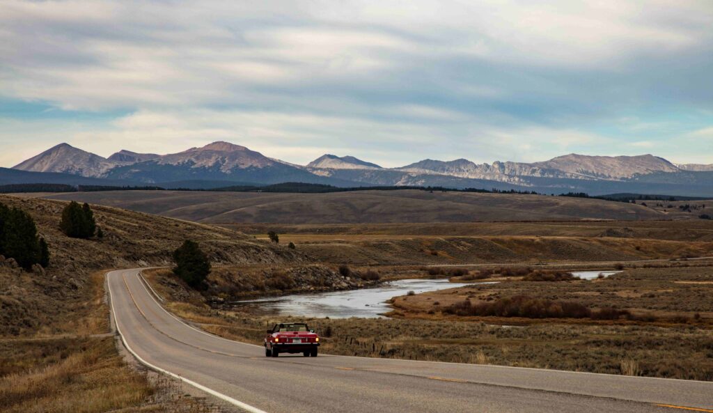 A classic red car drives into the distance with giant, dramatic, mountains in the horizon. Bright, sunny, beautiful day in Southwest Montana for a scenic drive. Driving near Dillon, Montana. Photo by Jim Ward.