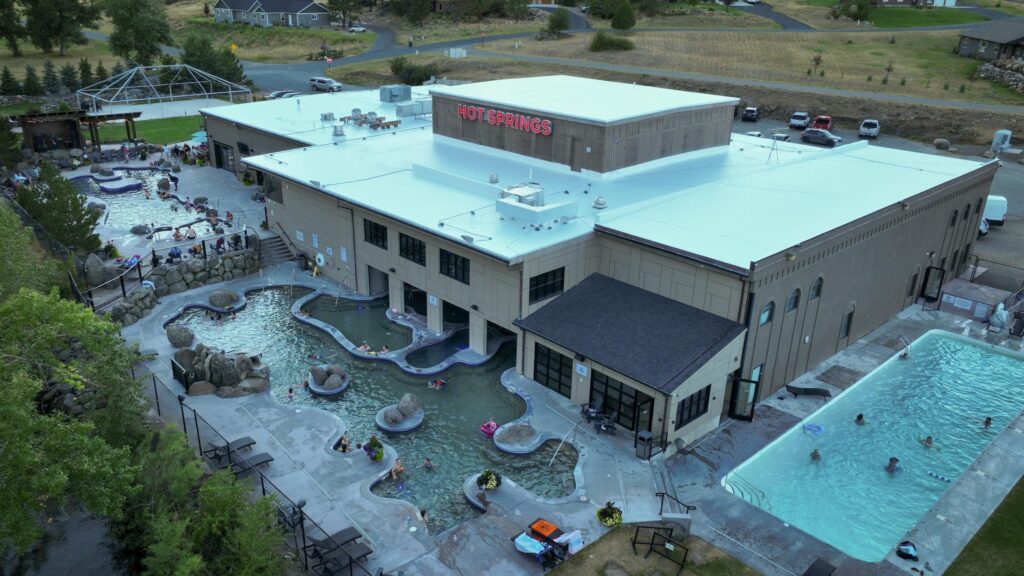 Aerial view of Broadwater Hot Springs in Helena, Montana, capturing the full scope of the facility's outdoor soaking pools, including several naturally shaped geothermal mineral pools with boulder accents, a large lap swimming pool, and a covered outdoor pavilion, all connected to the main indoor hot springs building. Guests enjoy the warm mineral waters across multiple pools as the residential and rural landscape of Helena surrounds the property, highlighting Broadwater Hot Springs as one of the most visited and beloved hot spring destinations in Southwest Montana. Photo by Sarah Bolt, Tempest Technologies.